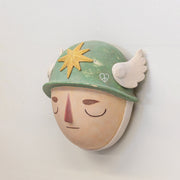 Sculpture of a person's head with a solemn yet sweet expression, They wear a military helmet with a star in front and angel wings on the side.