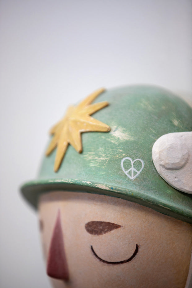 Sculpture of a person's head with a solemn yet sweet expression, They wear a military helmet with a star in front and angel wings on the side.