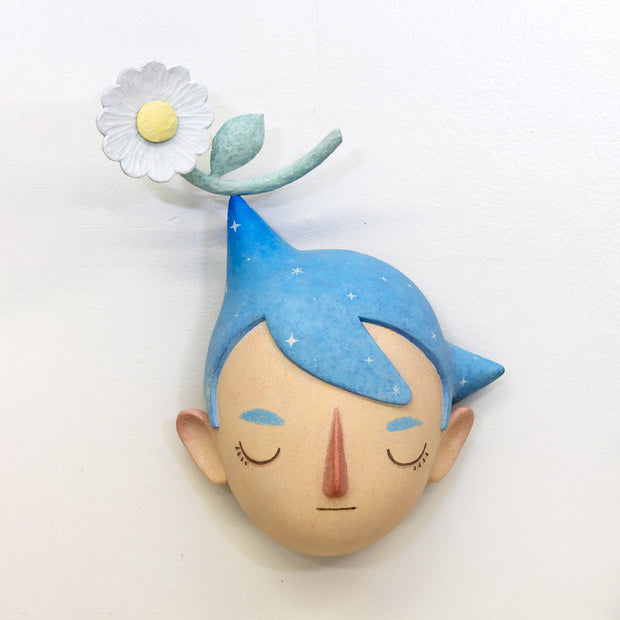 Sculpture of a person's head with a solemn yet sweet expression. They have pointed blue starry hair with a white flower sitting atop their head.