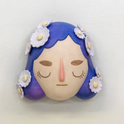 Sculpture of a person's head with a solemn yet sweet expression. They have mid length purple hair with white flowers all over like hair pins.