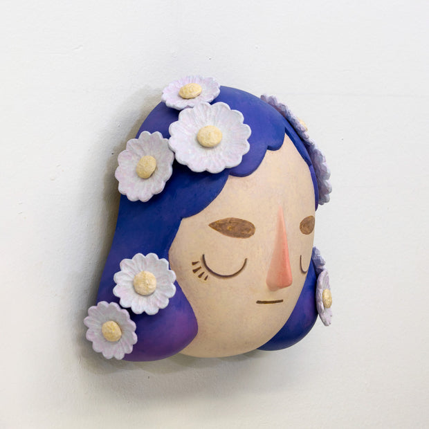Sculpture of a person's head with a solemn yet sweet expression. They have mid length purple hair with white flowers all over like hair pins.