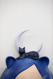 Sculpture of a person's head with a solemn yet sweet expression. They have mid length blue galactic pattern hair and small animal ears. A cat and a purple crescent moon sit atop their head.