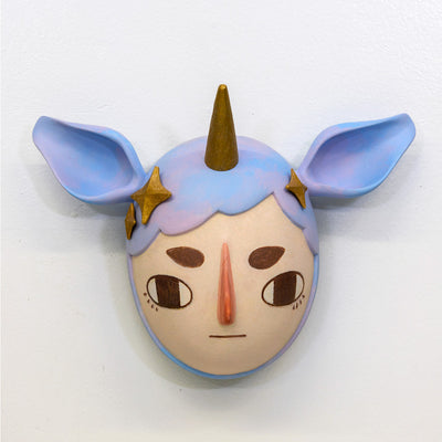 Sculpture of a person's head with a solemn yet sweet expression. They wear a hat that covers their hair shaped like a bluish purple unicorn, with ears and a golden horn. Golden stars decorate the sides like hair pins.