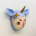 Sculpture of a person's head with a solemn yet sweet expression. They wear a hat that covers their hair shaped like a bluish purple unicorn, with ears and a golden horn. Golden stars decorate the sides like hair pins.