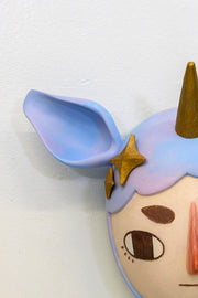 Sculpture of a person's head with a solemn yet sweet expression. They wear a hat that covers their hair shaped like a bluish purple unicorn, with ears and a golden horn. Golden stars decorate the sides like hair pins.
