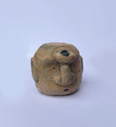 A hand-carved wood sculpture of a stylized face with a prominent bulbous nose and dark painted details, with a large 3rd eye atop its head, set against a plain light blue background.