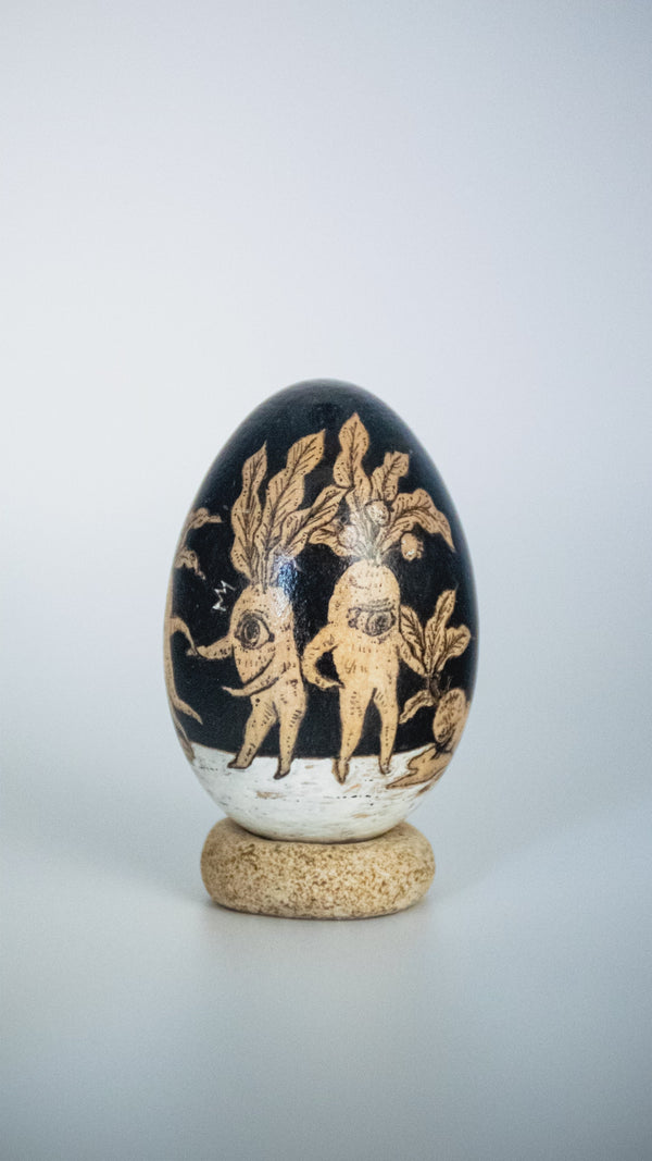 A decorative wooden egg with a glossy black ink illustration of botanical and animal motifs, resting on a small round cork base against a light gray background.