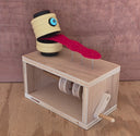 A wooden sculpture sitting atop a wooden box with a crank handle, featuring a yellow segmented head with a blue eye and a red tongue-like strip resting on metal pins, set against a wood-grain background.