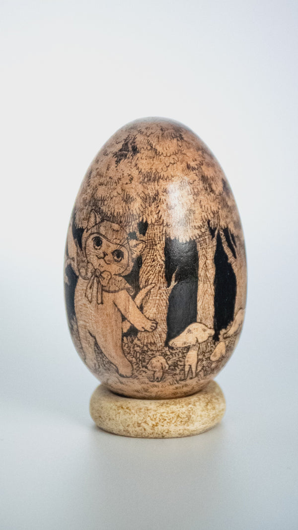 A carved wooden decorative egg sculpture with detailed hand-painted scenes of animals and trees, displayed on a small round wooden stand against a light gray background.