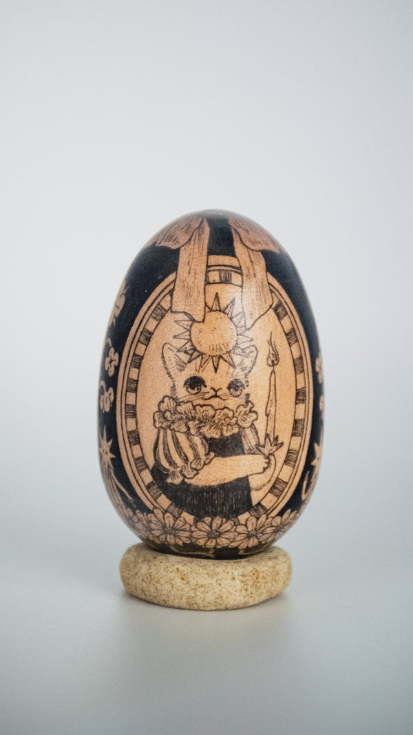 A hand-painted wooden egg with a detailed illustration of a cat holding a candle, with a sun overhead and within a nice border.