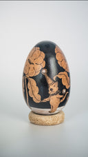 A decorative wooden egg with black illustrations of floral and animal illustrations, displayed upright on a small cork-like base against a light gray studio background.