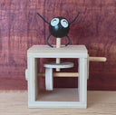 A wooden mechanical toy with a black round character figure on top, featuring white eyes and black thin arms and legs, set against a rustic reddish-brown wood background. Video shows the side lever being cranked.