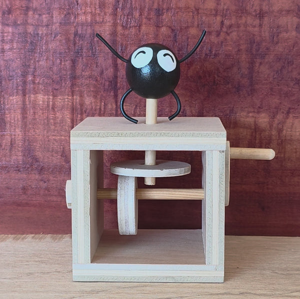 A wooden mechanical toy with a black round character figure on top, featuring white eyes and black thin arms and legs, set against a rustic reddish-brown wood background. Video shows the side lever being cranked.
