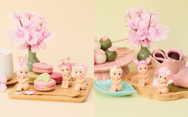 Promo photos of small Kewpie baby figures, sitting with their head in their hands and their elbows on their knees. They are all nude aside from pink or yellow hood hats of animals with cherry blossom decor.