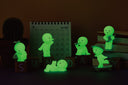 6 mini figures of Smiski characters, in various poses related to relaxation or fun activities. Options are: watering plant, blowing bubbles, sleeping w/ cat, flying a paper plane, skateboarding and playing ukelele. Glowing in the dark.