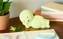 Light green cartoon-like creature, laying on its side with its hand propping up its head.  It sits on a desk with stationery items.