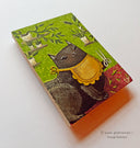 Painting on rectangular wood panel of a black cartoon cat wearing a yellow bib and holding shears, with several house plants on shelves behind.