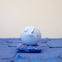 Small carved wooden sculpture of a blue sphere with a closed eye, calm smiling expression. It rests atop of a purple wooden pillow with splatter pattern, carved to look soft.