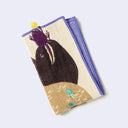 Rectangular cream colored towel with a purple outline and imagery of a brown stream running past oval shaped brown rocks. Small blue tadpoles swim in the stream and a bigger purple tadpole is atop a large rock, holding a dandelion.