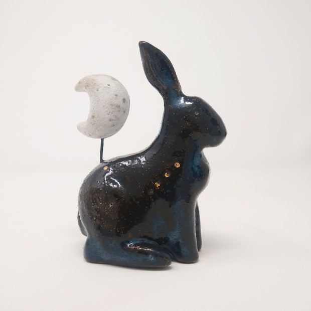 Ceramic sculpture of a black and blue bunny without any facial features and simplistic body features, sitting on its hind legs. Atop its back is a white speckled crescent moon.