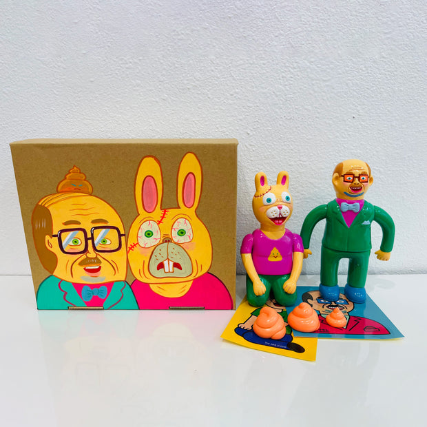 2 soft vinyl figures, a man with glasses dressed in a green suit and a yellow rabbit with a human body, sitting on its knees. 3 small orange poop swirls are in front of them. All stand next to a painted box.