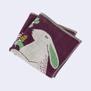 Reddish purple square handkerchief featuring an illustration of a light gray hare with green inner ears holding a small plant. It stands on its hind legs next to a large plant, the same size as itself.