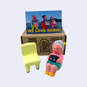 Vinyl figure of an old woman sitting with a radio on her lap. She sits on the ground, next to a light neon green chair. Behind the figures is their product packaging and a postcard.