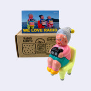 Vinyl figure of an old woman sitting with a radio on her lap. She sits on a bright pastel green chair. Behind the figures is their product packaging and a postcard.