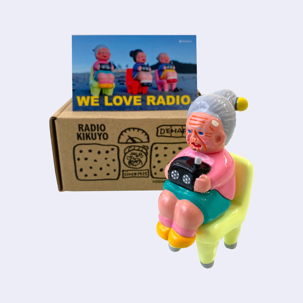 Vinyl figure of an old woman sitting with a radio on her lap. She sits on a bright pastel green chair. Behind the figures is their product packaging and a postcard.