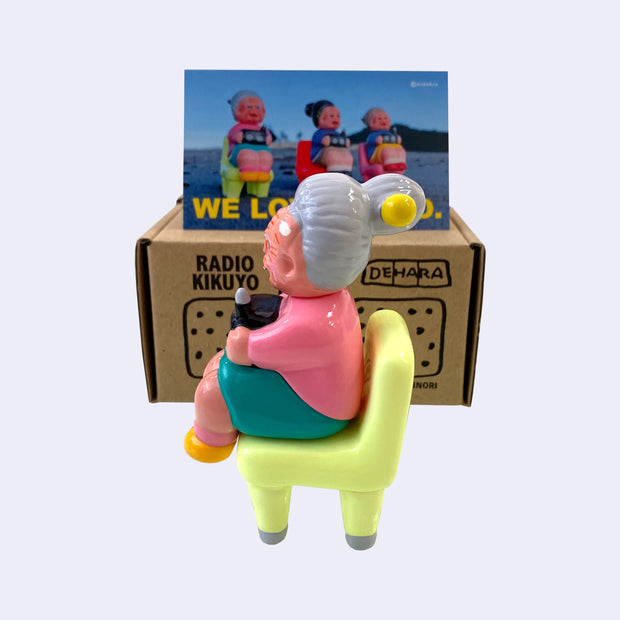Vinyl figure of an old woman sitting with a radio on her lap. She sits on a bright pastel green chair. Behind the figures is their product packaging and a postcard.