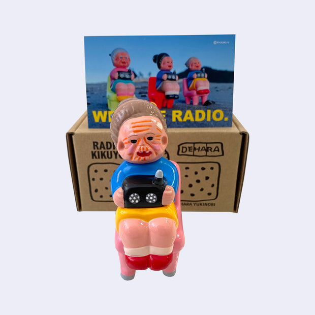 Vinyl figure of an old woman sitting with a radio on her lap. She sits on a pink chair. Behind the figures is their product packaging and a postcard.