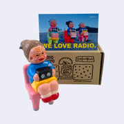 Vinyl figure of an old woman sitting with a radio on her lap. She sits on a pink chair. Behind the figures is their product packaging and a postcard.