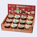Ramen toppings cultivation kit in a cardboard display box, showing multiple small bowls of ramen toppings with wooden sticks and illustrated plant pots on the red inner lid.