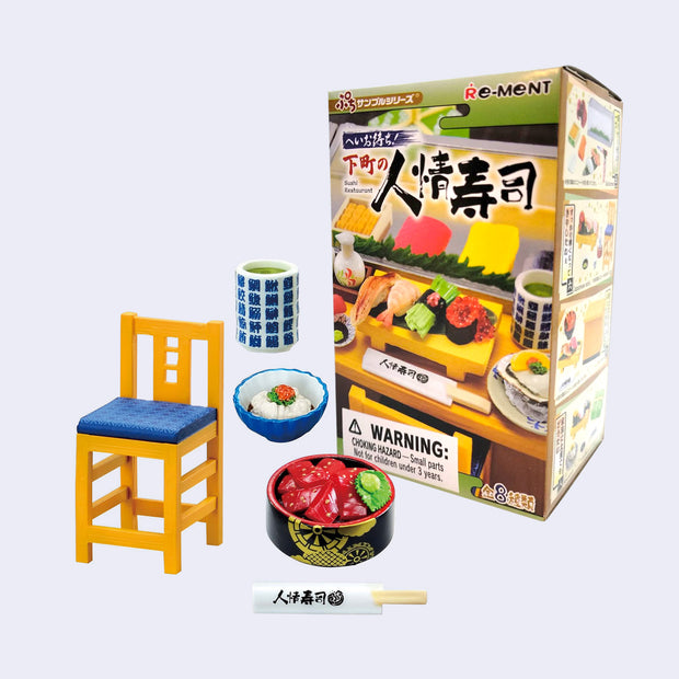 Miniature dining set with chair, tableware, chopsticks and plates of sushi. They are positioned in front of their blind box packaging.