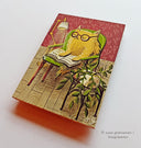 Painting on wood of a fluffy yellow cartoon cat sitting in a green armchair and reading a book, with a lamp shining down onto it. A houseplant is on a side table nearby.