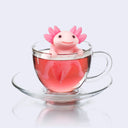 Pink silicone tea infuser shaped like an axolotl with a smiling face, sitting in a glass mug.. Its hands hang over the rim of the cup to keep itself up and tea diffuses from small holes in its body.