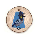 Wooden disc with a painting of a black bird with 3 yellow eyes and a hat.