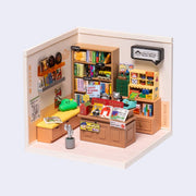 3D assembled cube diorama of a modern and stylish bookstore, with bookcases and racks filled with different colored covered books and magazines. There is a small reading bench that has cute pillows and signage around the store. 2 sides have wall backings and 2 sides are open.