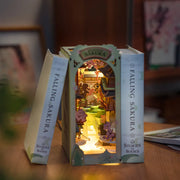 Fully assembled book nook diorama, featuring a small girl looking into a house with sakura blossoms falling around the scene. Bookend is lit up and nestled between 2 books.