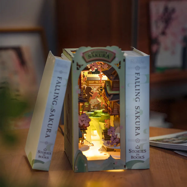Fully assembled book nook diorama, featuring a small girl looking into a house with sakura blossoms falling around the scene. Bookend is lit up and nestled between 2 books.