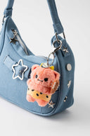 Plush keychain of a fluffy pinkish orange monster with tiny yellow horns and tiny fangs. It wears a cheetah skin loin cloth and holds a spiked club. It hangs from a blue handlebag.