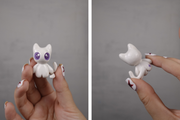 White and purple cat ceramic figure held by a hand against a gray background