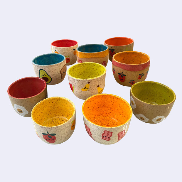10 ceramic bowls with bright insides of all different colors, including yellow, orange, green, blue, or pink/red. Exterior of bowls have little drawings of foods, such as fruits or breakfast items.