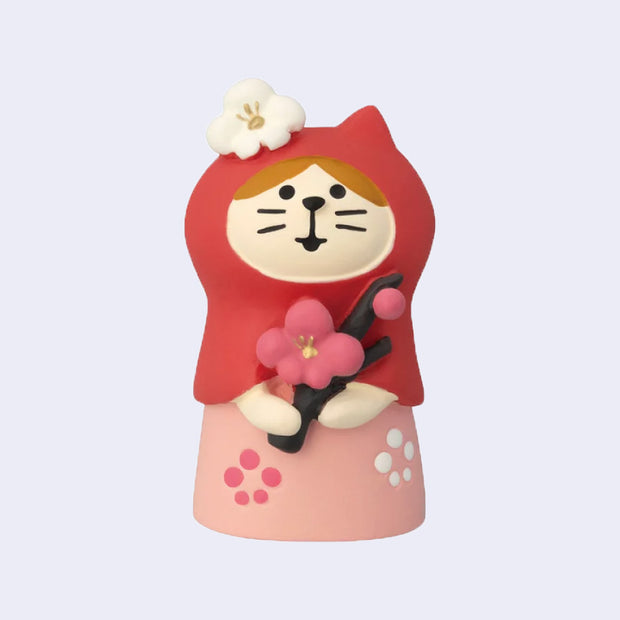 Small figure of a cat with a red hood and pink dress holding a branch with a cherry blossom on it.