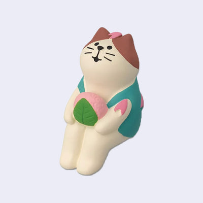 Sitting figure of a white cat with brown ears, wearing a teal vest and holding a sakura mochi with cherry blossom petals on its head and body.