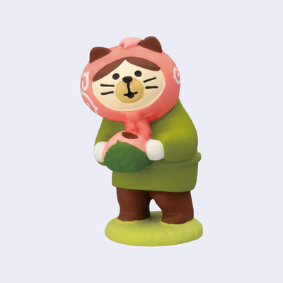 Small figure of a white and brown cat wearing green and brown clothes and a pink head scarf. It stands on a green mound and holds a sakura mochi.