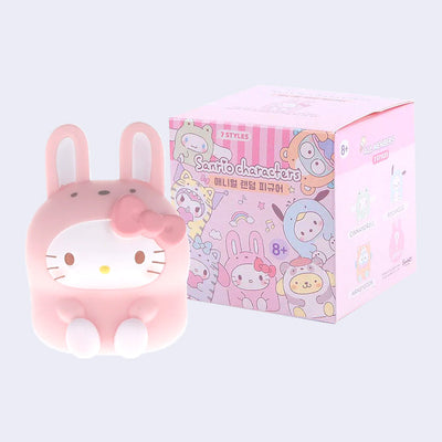 Pink Hello Kitty figurine wearing a costume and bunny ears, sitting in front of a box of Sanrio character figurines.