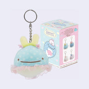 Plush keychain of a cute cartoon whale with bunny ears and a tutu, with its product packaging behind it.