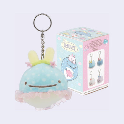 Plush keychain of a cute cartoon whale with bunny ears and a tutu, with its product packaging behind it.