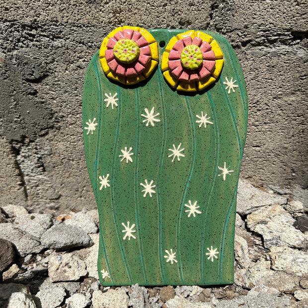Flat ceramic cactus sculpture, green with white star shaped bursts meant to replicate spikes. Atop are 2 blooming flowers, yellow and pink with yellow centers.